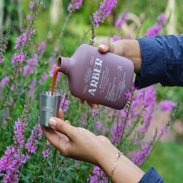 Hands pour Arber Organic Bio Fungicide from a matte mauve bottle into a silver measuring cup, surrounded by purple flowers.