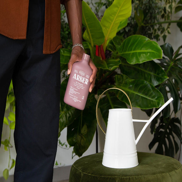Person holding a mauve Arber Organic Bio Fungicide bottle next to a white watering can and green plants.