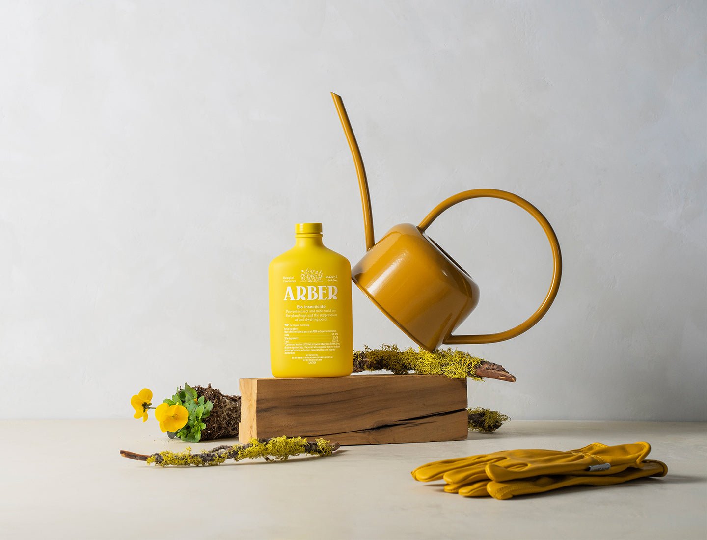 Bright yellow Arber Organic Insecticide bottle, matching watering can, and gardening gloves on a wooden block with moss and yellow flowers.