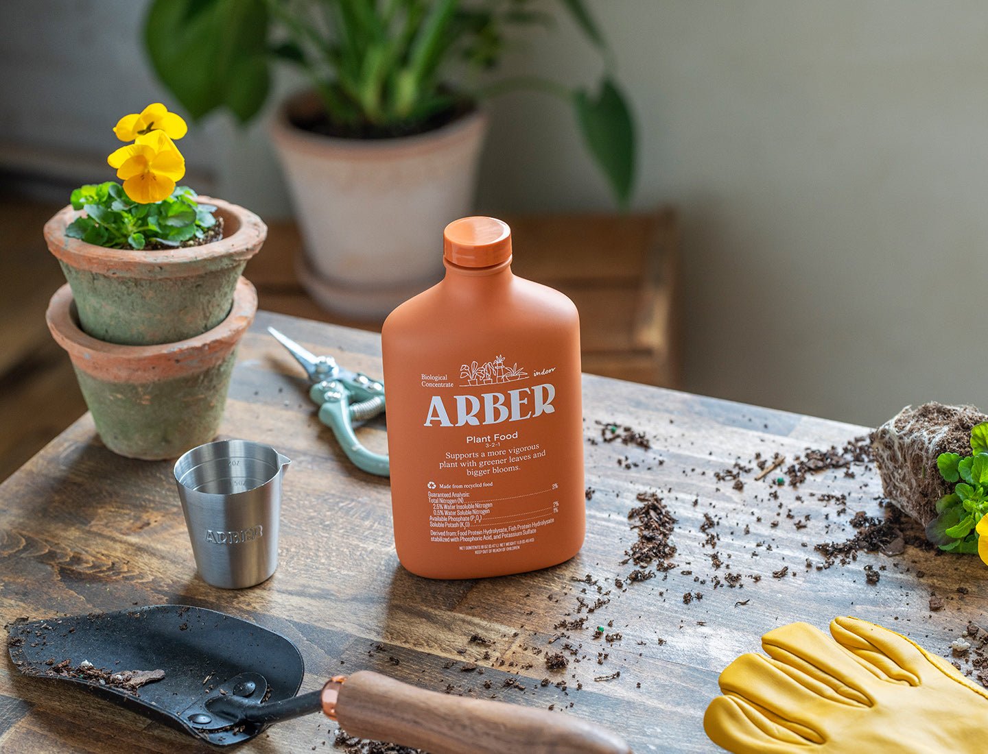 Orange Arber Organic Plant Food 16oz bottle, measuring cup, gardening tools, and potted plants on a wooden table.