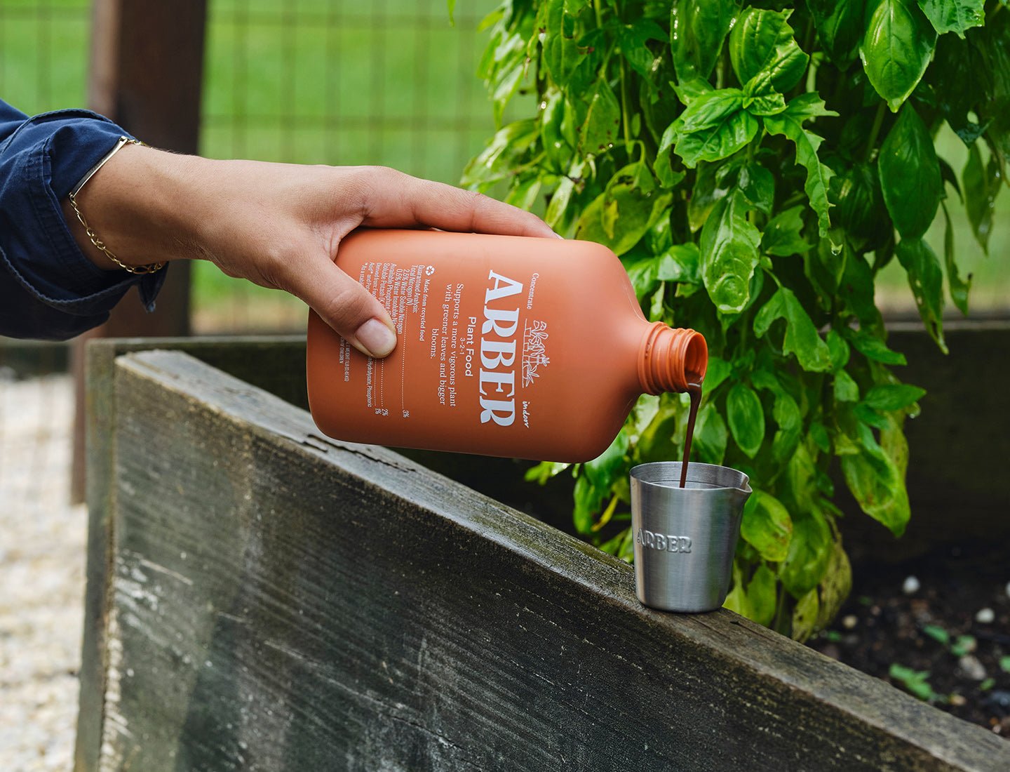 Hand pouring Arber Organic Plant Food from a 16oz terracotta bottle into a silver measuring cup over a raised garden bed.