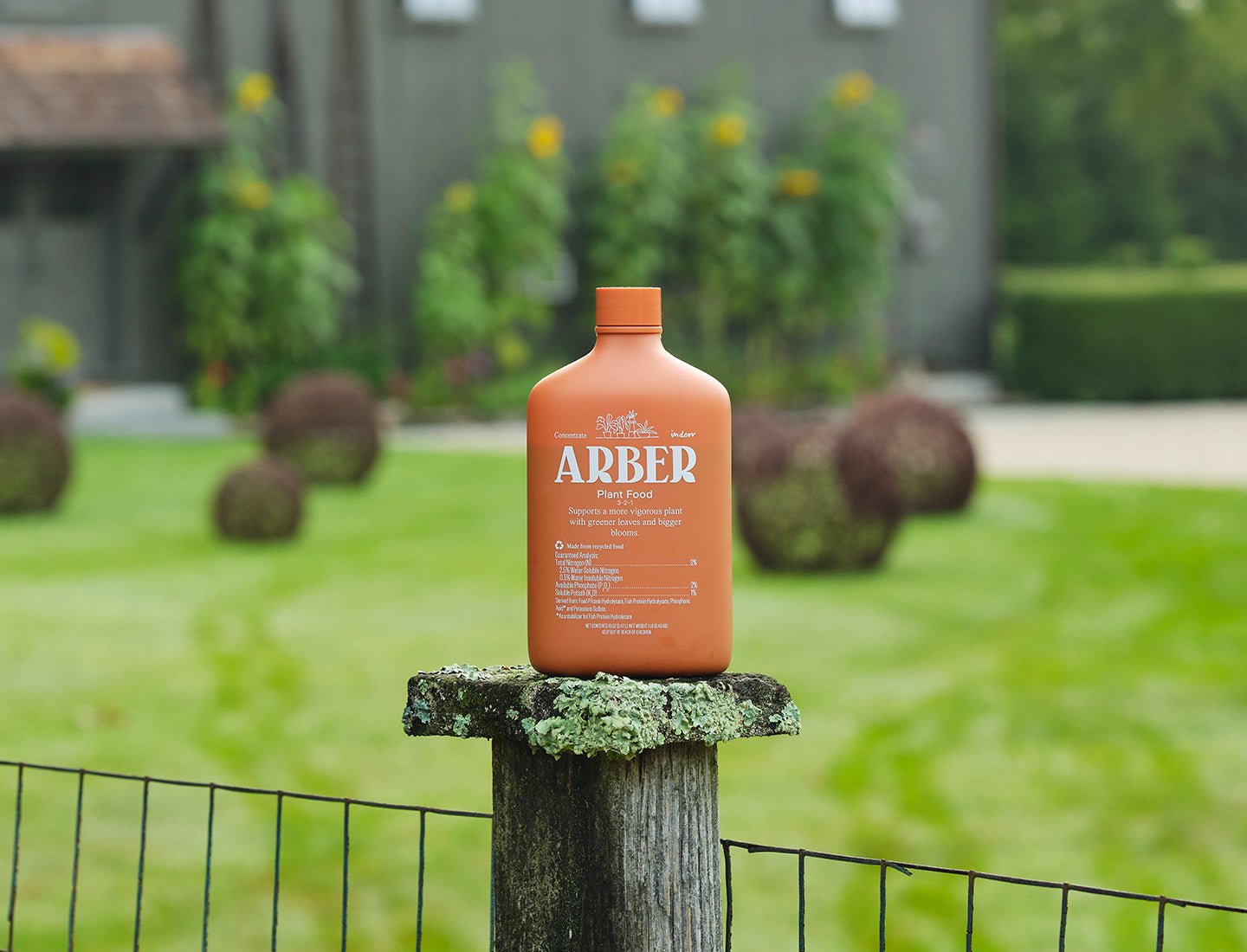 Orange 16oz Arber Organic Plant Food bottle with white label, on a mossy wooden post in a garden.