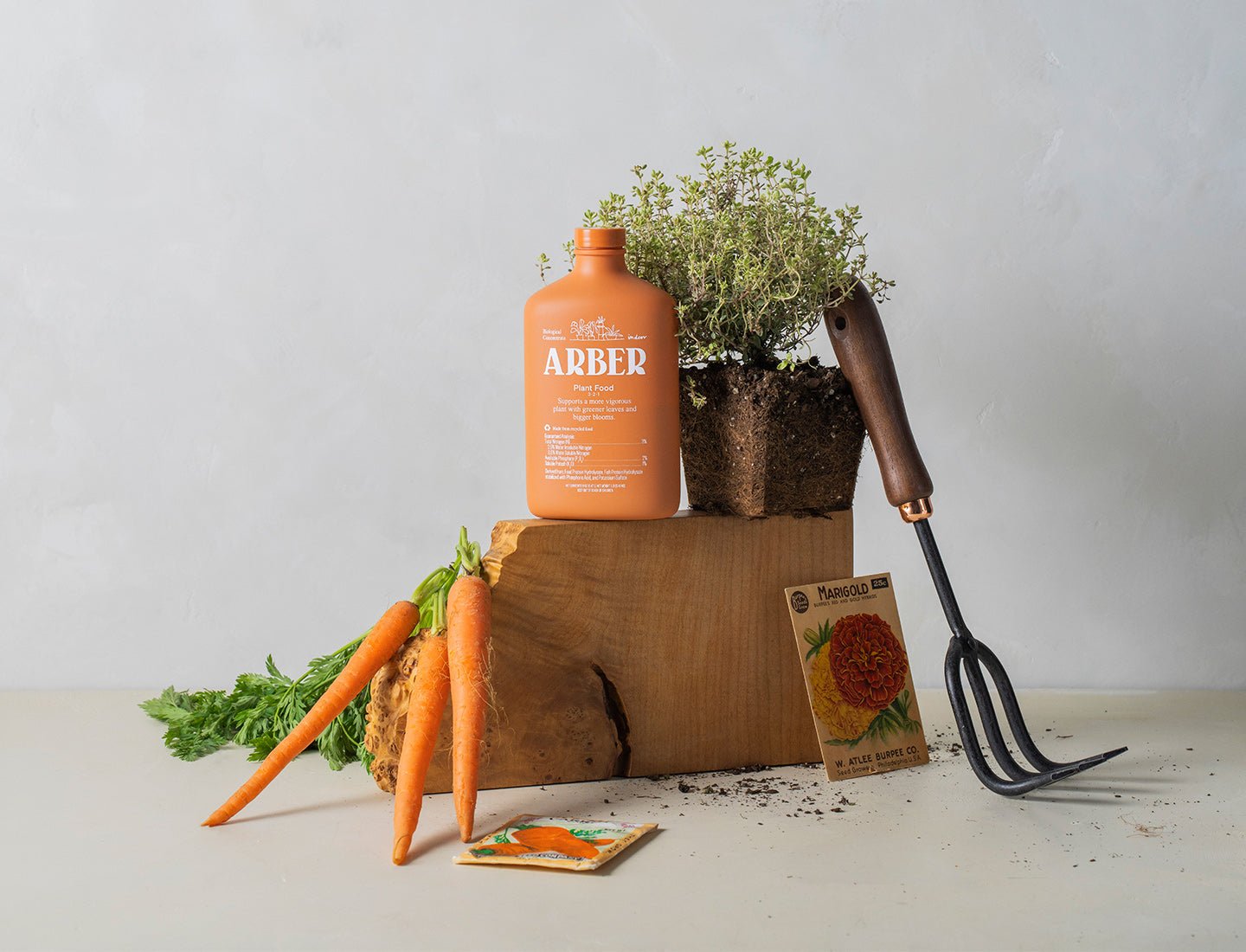 Orange Arber Plant Food bottle on wood block with carrots, thyme plant, garden fork, and seed packets.